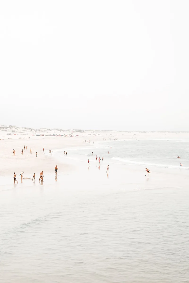 Aerial beach photo of people walking and playing along a soft pastel shoreline with gentle waves.