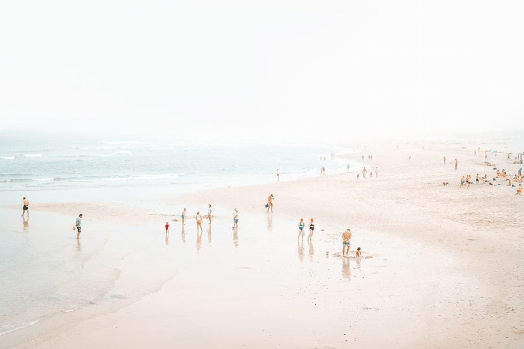 Aerial beach photograph with families and beachgoers scattered across soft wet sand near gentle surf.