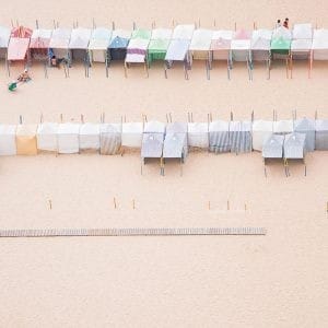 Rows of pastel beach huts lined up on a sandy beach, captured from above