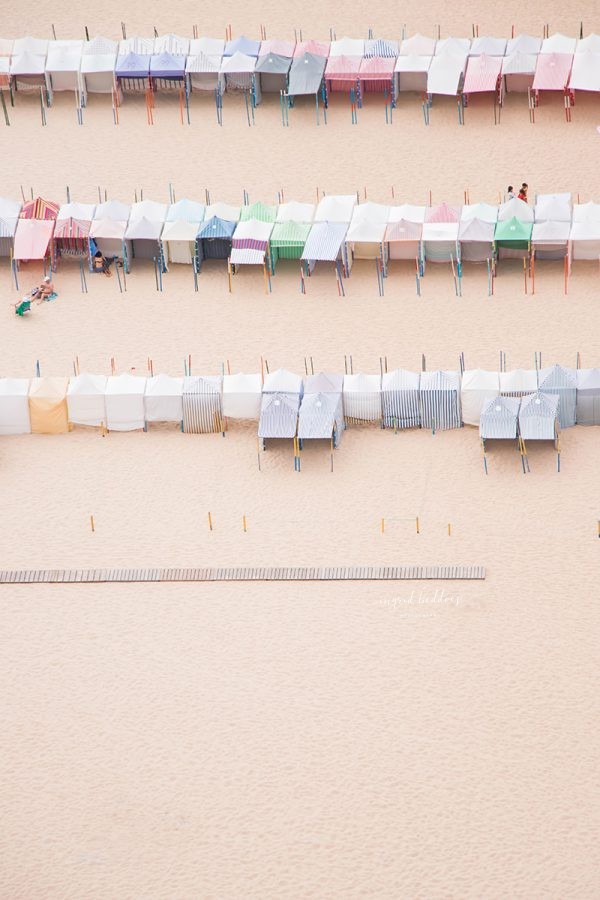 Rows of pastel beach huts lined up on a sandy beach, captured from above