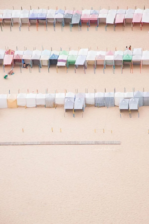 Rows of pastel beach huts lined up on a sandy beach, captured from above