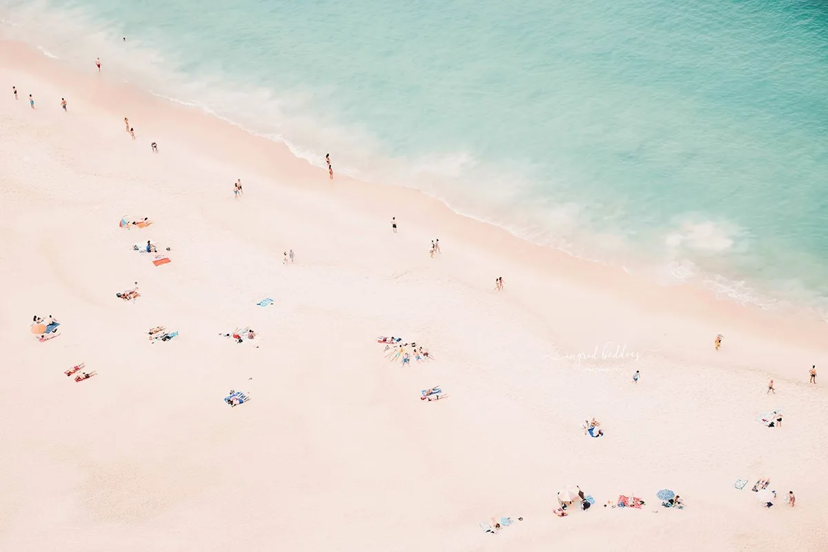 Aerial shot of people scattered along a peaceful, pastel-toned beach and aqua sea