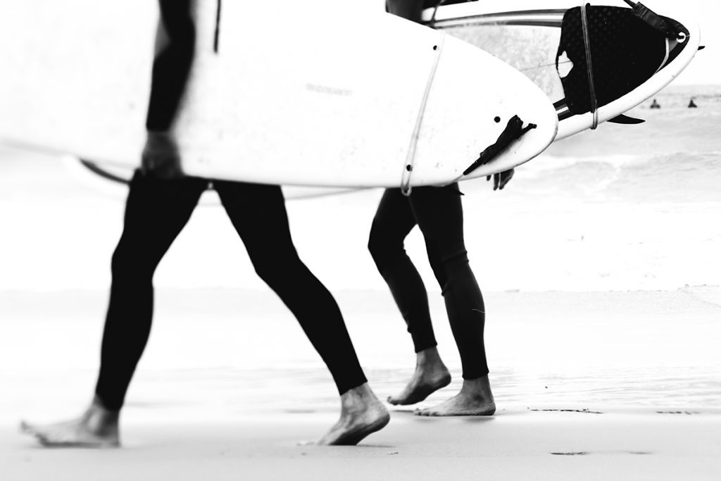 Black and white photo of surfers walking barefoot on the sand carrying surfboards.