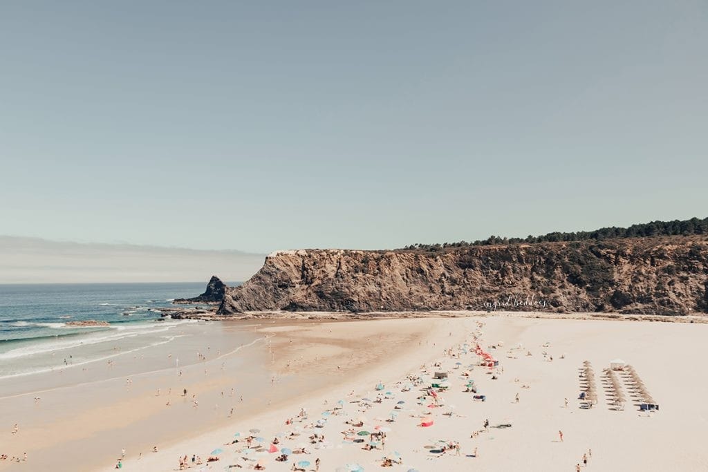 Wide view of a beach with sunbathers, colorful umbrellas, and dramatic cliffs meeting the sea.
