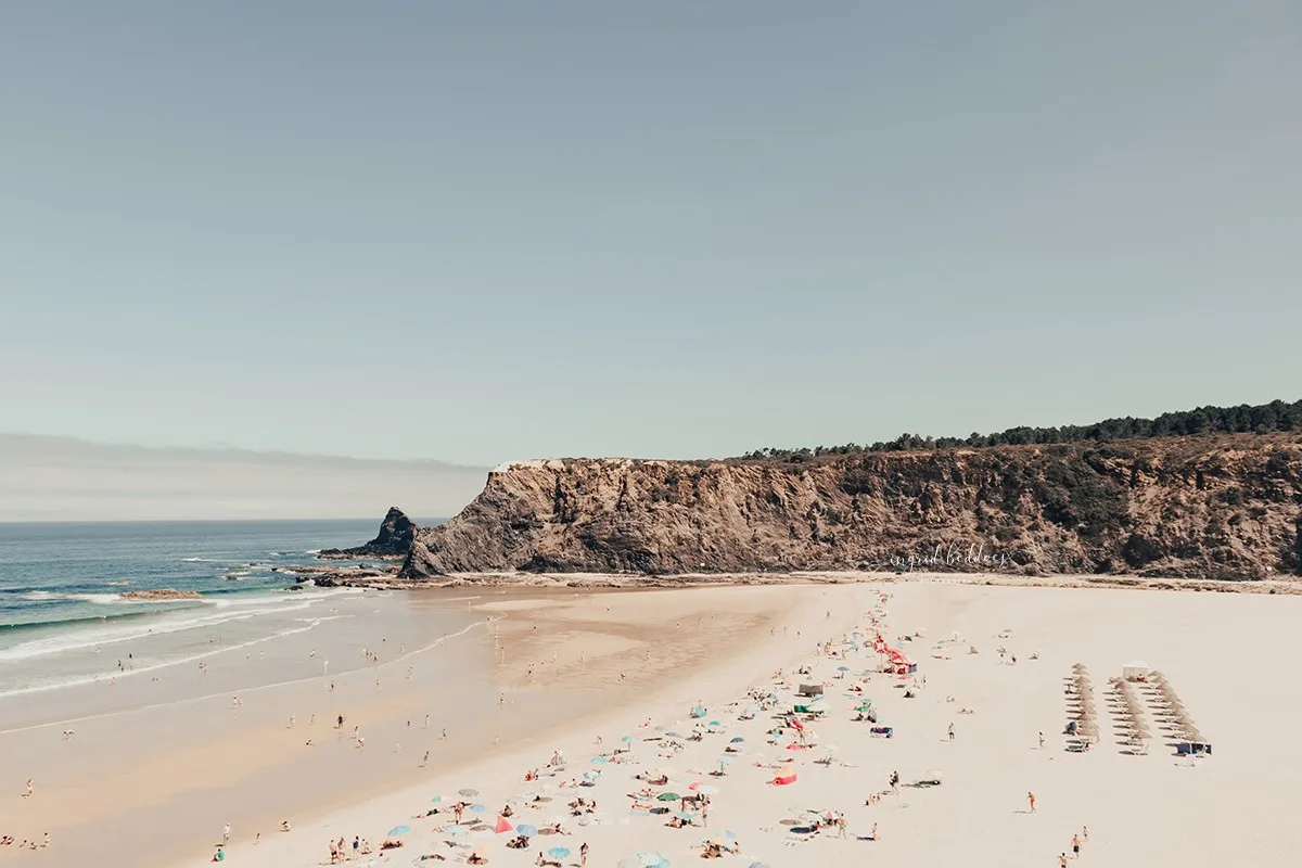 Wide view of a beach with sunbathers, colorful umbrellas, and dramatic cliffs meeting the sea.